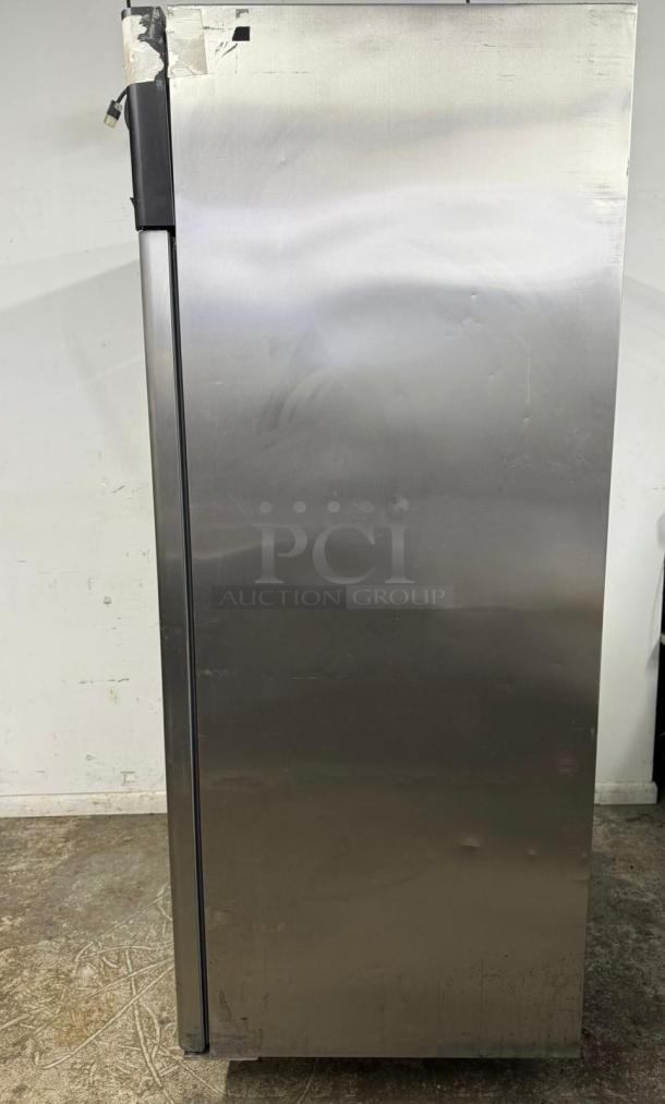 Stainless steel commercial refrigerator, showing wear with surface dents and tape residue, left side view.