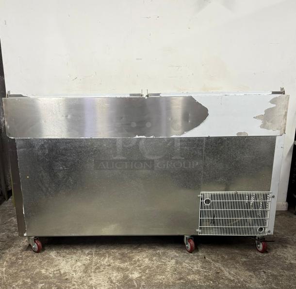 Stainless steel commercial refrigerator on wheels, scratched surface, visible wear. Ventilation grille on lower right.