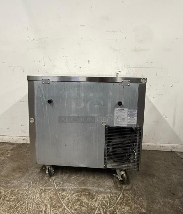 Commercial stainless steel refrigerator unit on wheels, visible scuffs, ventilation panel, and caution labels.