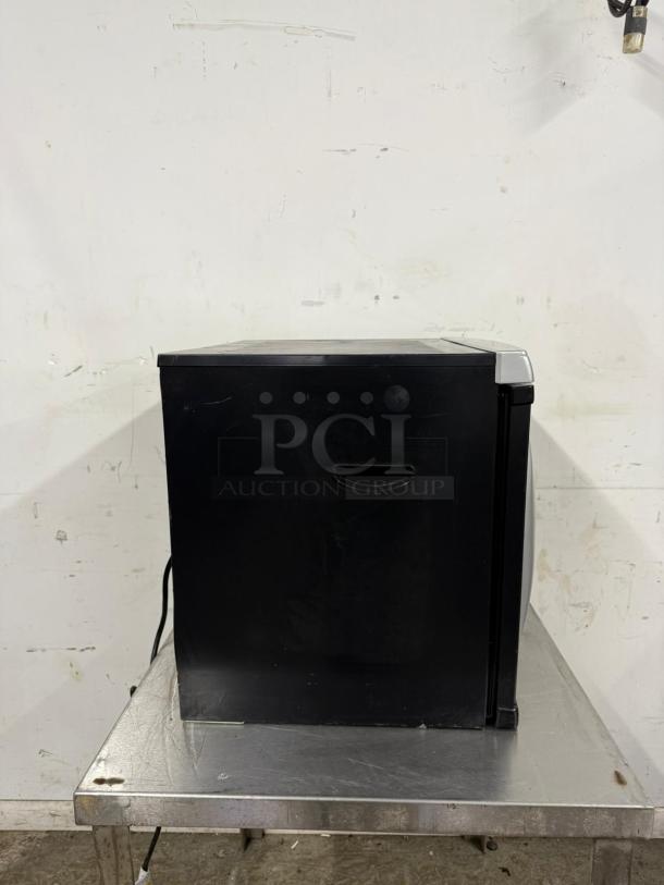 Black mini fridge on a metal table, showing side view with a handle. Slightly scratched surface, plug visible.