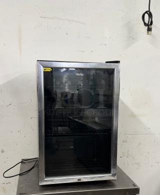 Haier mini fridge with glass door, black interior, shelving. Used condition, visible scratches. Energy efficiency label visible.