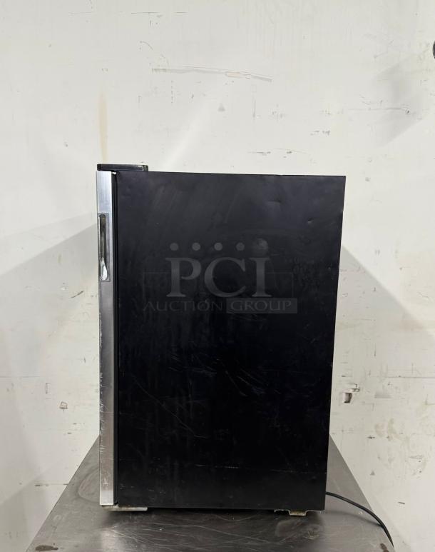 Black mini refrigerator with a stainless steel handle, visible wear and surface scratches, resting on a metal surface.