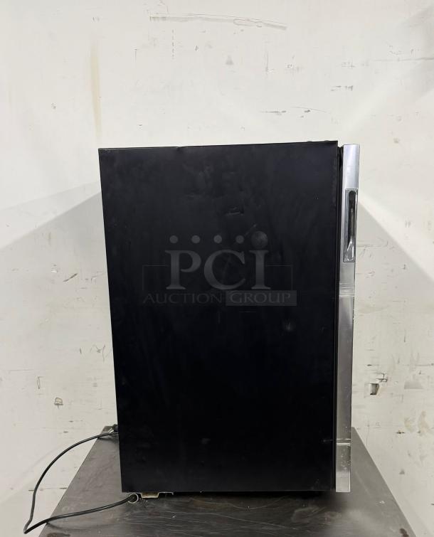 Black mini fridge with sleek stainless steel handle. Appears in good condition, minor surface scratches visible. Power cord attached.