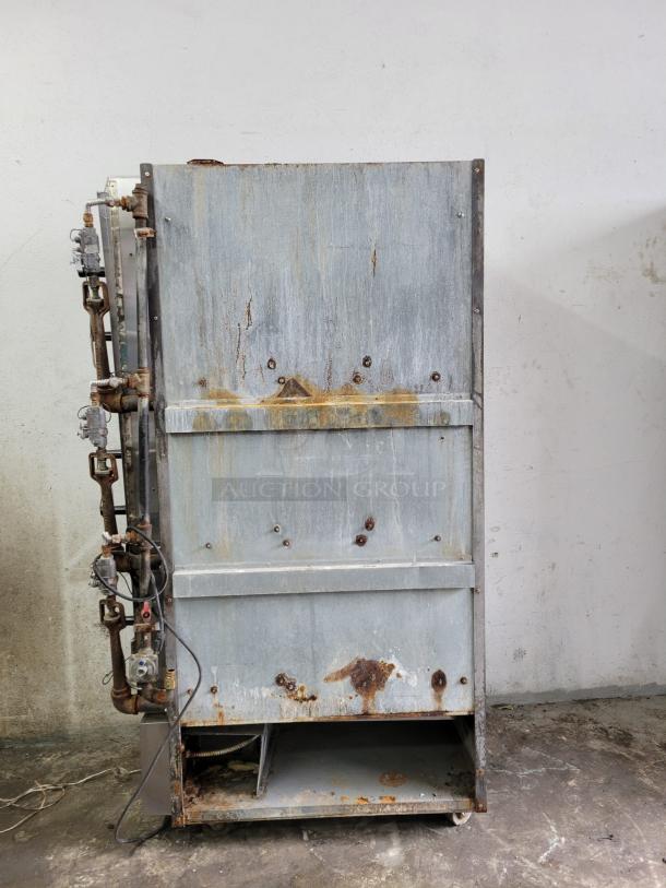 Large industrial metal furnace with visible rust and wear, located indoors. Shows attached pipes and wires. No visible labels.