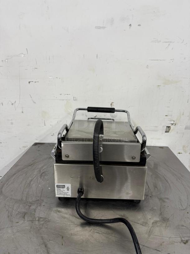 Commercial panini press in stainless steel, lightly used. Features include a heat-resistant handle and label indicating electrical specifications and safety warnings.
