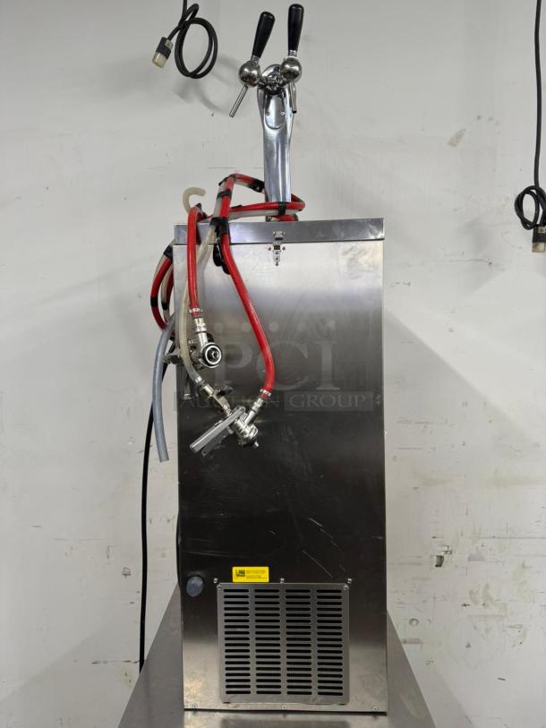 Commercial beer dispenser with dual taps and red hoses, stainless steel. Visible wear on surface. Yellow warning label below.