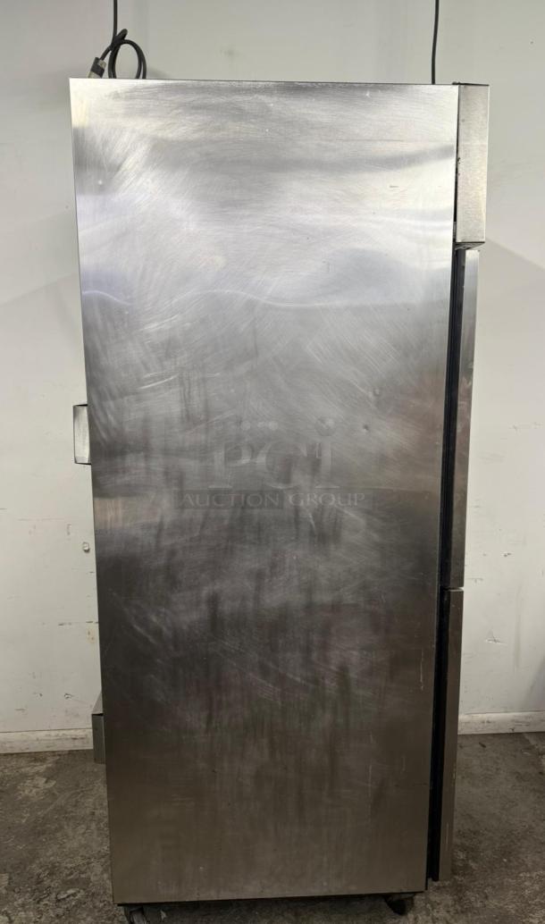 Commercial stainless steel refrigerator, upright design, visible wear and scratches, caster wheels for mobility. Plug on top.