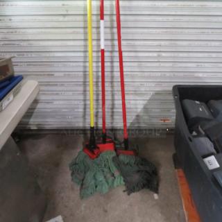 Assorted mops with colored handles in used condition, leaning against a metal backdrop.