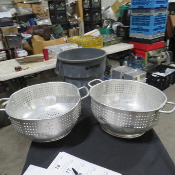 Two aluminum colanders with dual handles, perforated design, and a shiny finish, placed on a black surface.