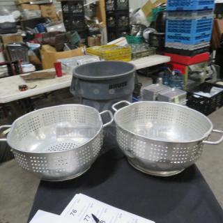 Two aluminum colanders with dual handles, perforated design, and a shiny finish, placed on a black surface.