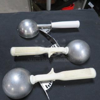Three white handle dishers on black surface, with stainless steel scoops. Good condition, suitable for portioning tasks.