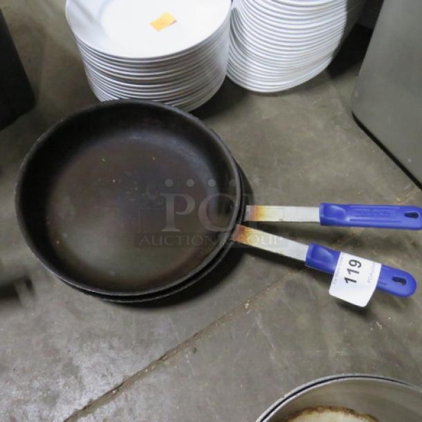 Assorted sauté pans with blue handles, signs of wear, and auction tag "119," displayed on a concrete floor.