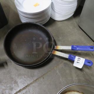 Assorted sauté pans with blue handles, signs of wear, and auction tag "119," displayed on a concrete floor.