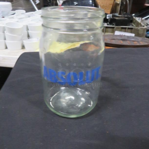 Clear glass mason jar labeled "ABSOLUT" in blue text, clean, no visible damage, background with stacked utensils.