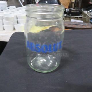 Clear glass mason jar labeled "ABSOLUT" in blue text, clean, no visible damage, background with stacked utensils.