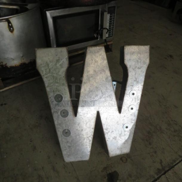 Aluminum letter shaped as M or W, metallic finish, industrial style, with bolt details, in used condition.
