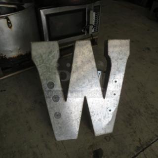 Aluminum letter shaped as M or W, metallic finish, industrial style, with bolt details, in used condition.