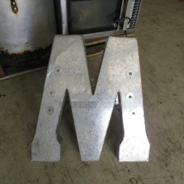 Aluminum letter "M" or "W," metallic finish, visible mounting hardware, rustic condition, no visible markings.