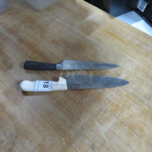 Two assorted chef knives on a wooden surface, featuring black and white handles; one knife labeled "18."