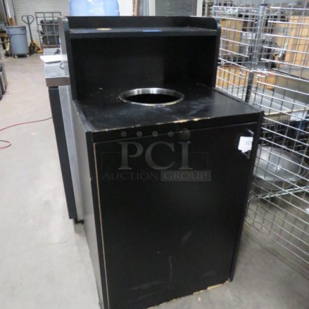 One-door trash receptacle with overshelf, 26x26x48 inches, black finish, visible wear on edges, round disposal opening.