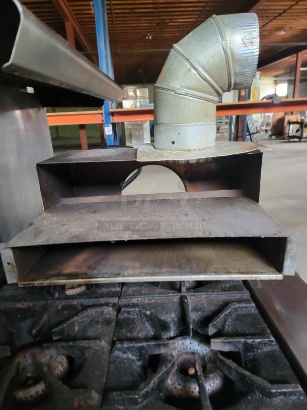 Commercial stove with metal flue duct, showing rust and wear. Features multiple burners and a stainless steel shelf.