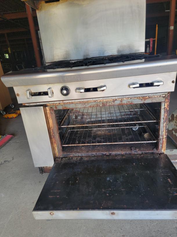 Commercial stainless steel stove with open oven door, showing rust and wear. Three control knobs; used condition.