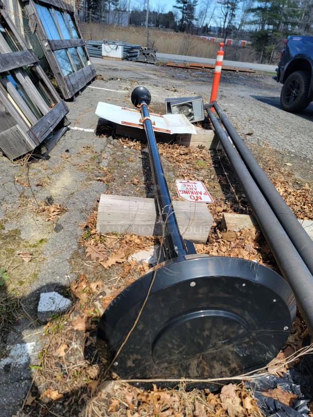 Black streetlamp post with round base, removed and lying on ground. Includes attached metal signs and nearby construction cones.