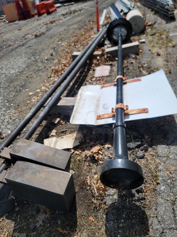 Industrial metal pole with mounting brackets, lying on gravel. Rust visible on brackets, indicating wear.