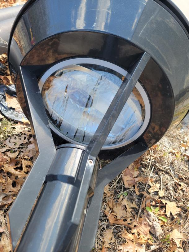 Outdoor floodlight with glass lens, black metal housing, slight cosmetic scratches, resting on leaf-covered ground.