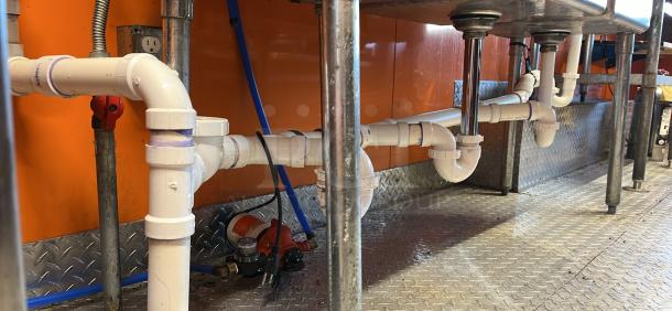 Plumbing under the sink of a 2014 Quality Trailers mobile kitchen, showing PVC pipes and a new water pump.
