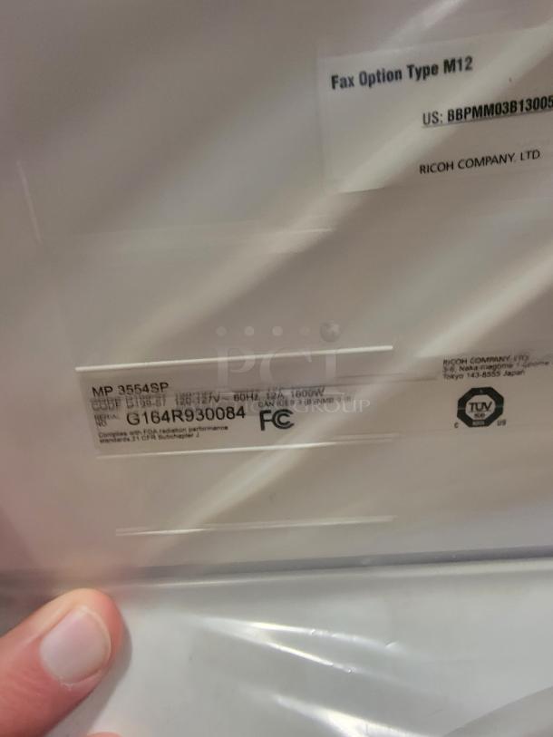 Ricoh MP 3554SP multifunction printer label showing model, code, and compliance marks. Finger holding the device in view.