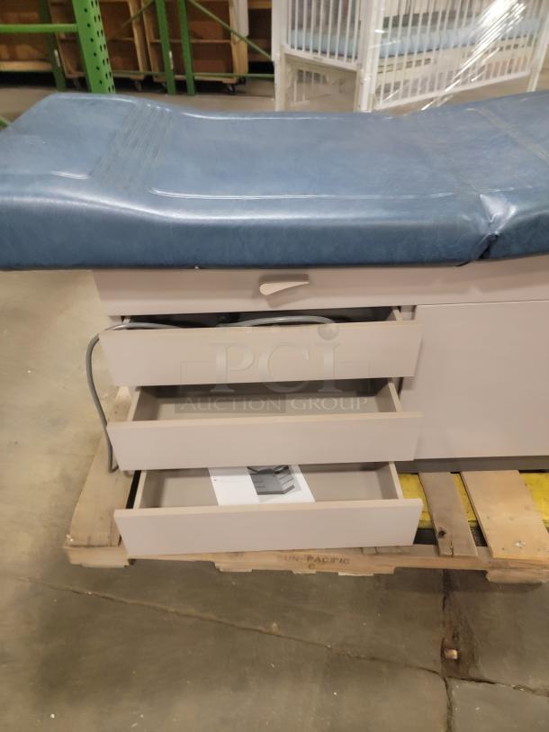 Ritter 104 examination table with blue padded surface, open storage drawers, and manual. Ideal for versatile uses.