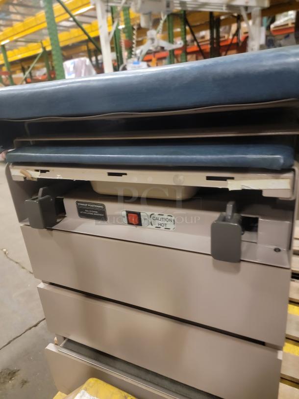 Ritter 104 Examination Table with blue cushioned top. Features include storage drawers, sturdy design, and caution hot markings.