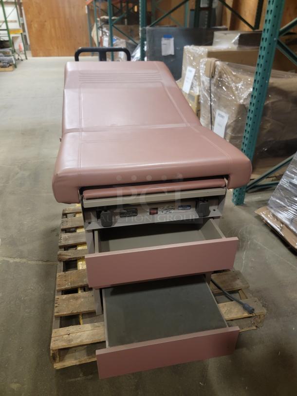 Ritter 104 Examination Table in pink, on a wooden pallet. Features multiple storage drawers, sturdy design, good condition.