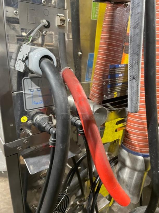 Industrial machine with connected hoses and cables, metallic casing, visible labels indicating technical specifications.