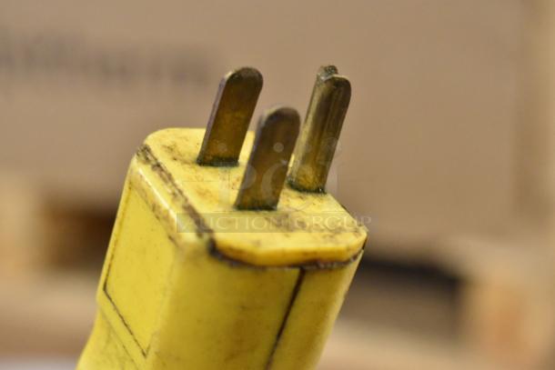 Close-up of a yellow electrical plug with three metal prongs, showing signs of wear and slight discoloration.