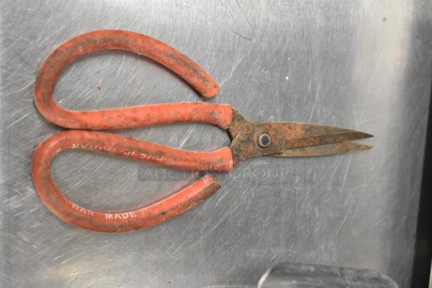 Rusty scissors with red handles, labeled "Made in Taiwan," placed on a metal surface.