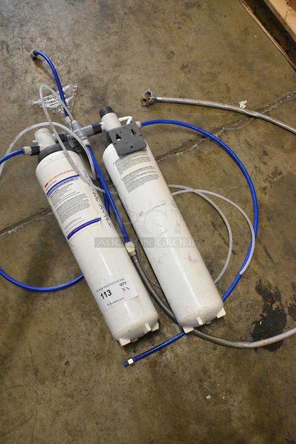 Two 3M water filter cartridges with attached hoses, seen on a concrete floor. Labels show specifications and auction details.