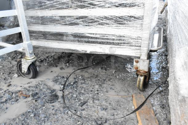 Two metal commercial pan transport racks with baking pans, wrapped in protective material, shown on a wet, gravel surface.