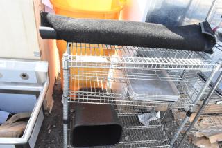 Chrome finish 3-tier wire shelving unit containing a rolled floor mat and trays. Buyer must dismantle for shipping.