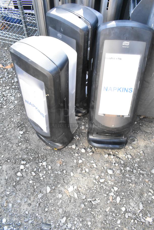 Three black and white napkin dispensers labeled "Tork Xpressnap" standing on gravel, part of auction lot with gray trash can.