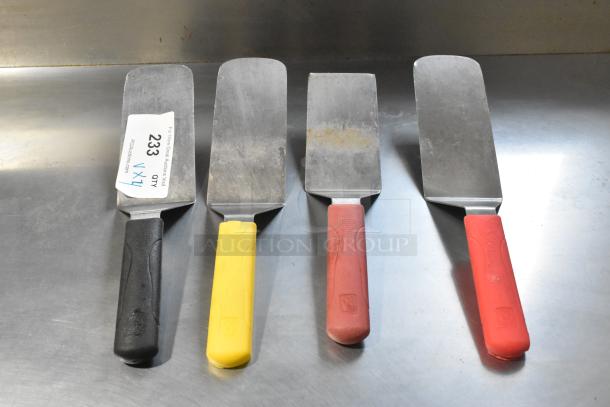 Four metal spatulas with colored handles: black, yellow, brown, and red. Slight wear visible. Label on one indicates quantity.