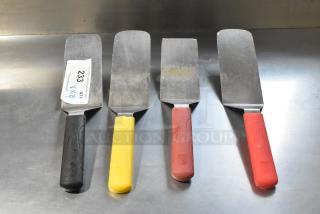 Four metal spatulas with colored handles: black, yellow, brown, and red. Slight wear visible. Label on one indicates quantity.