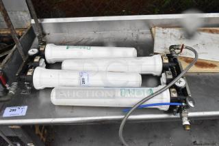 Industrial water filtration system with four white filter cartridges, pressure gauges, and attached hoses on a metal surface.