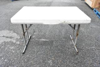 White folding table with metal legs, showing signs of wear and rust. Ideal for outdoor use or temporary setups.