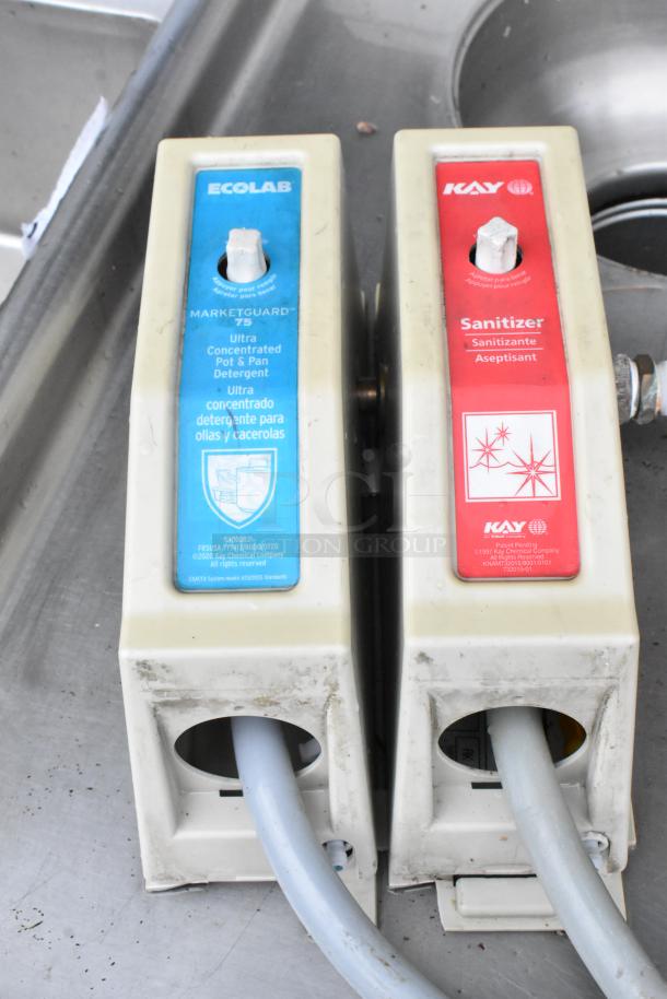 Ecolab detergent dispenser and Kay sanitizer dispenser on stainless steel sink. Visible wear on dispensers.