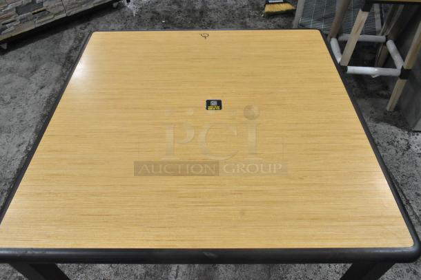 Square dining height table with woodgrain top and black metal edges. Four sturdy legs. Minor wear visible. Black label on top.