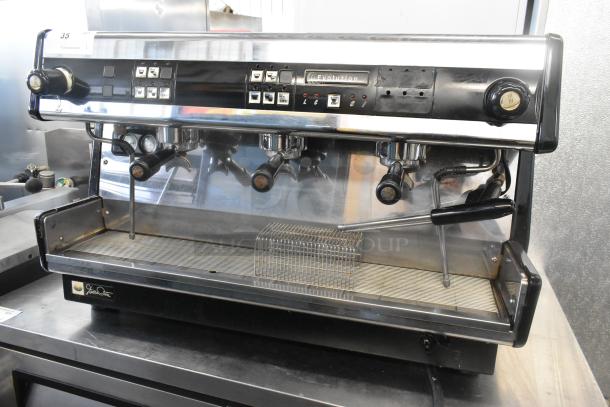 Dalla Corte stainless steel 3-group commercial espresso machine with three portafilters and two steam wands, featuring control buttons.