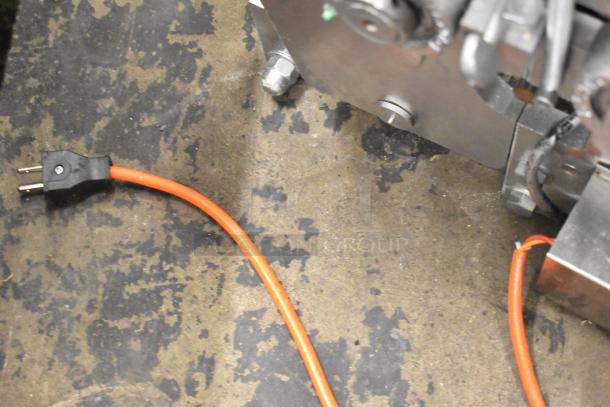 Metal countertop sheeter with orange power cord on textured surface, showing industrial design and connection details.