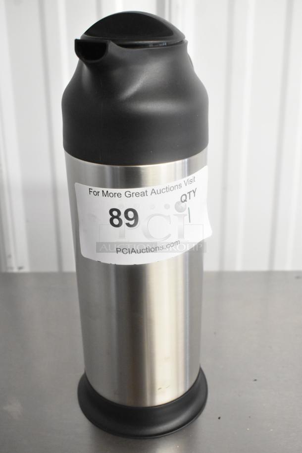 Stainless steel pitcher with black plastic lid, slight dents visible. Auction label: "For More Great Auctions Visit PCIAuctions.com, 89, QTY 1".
